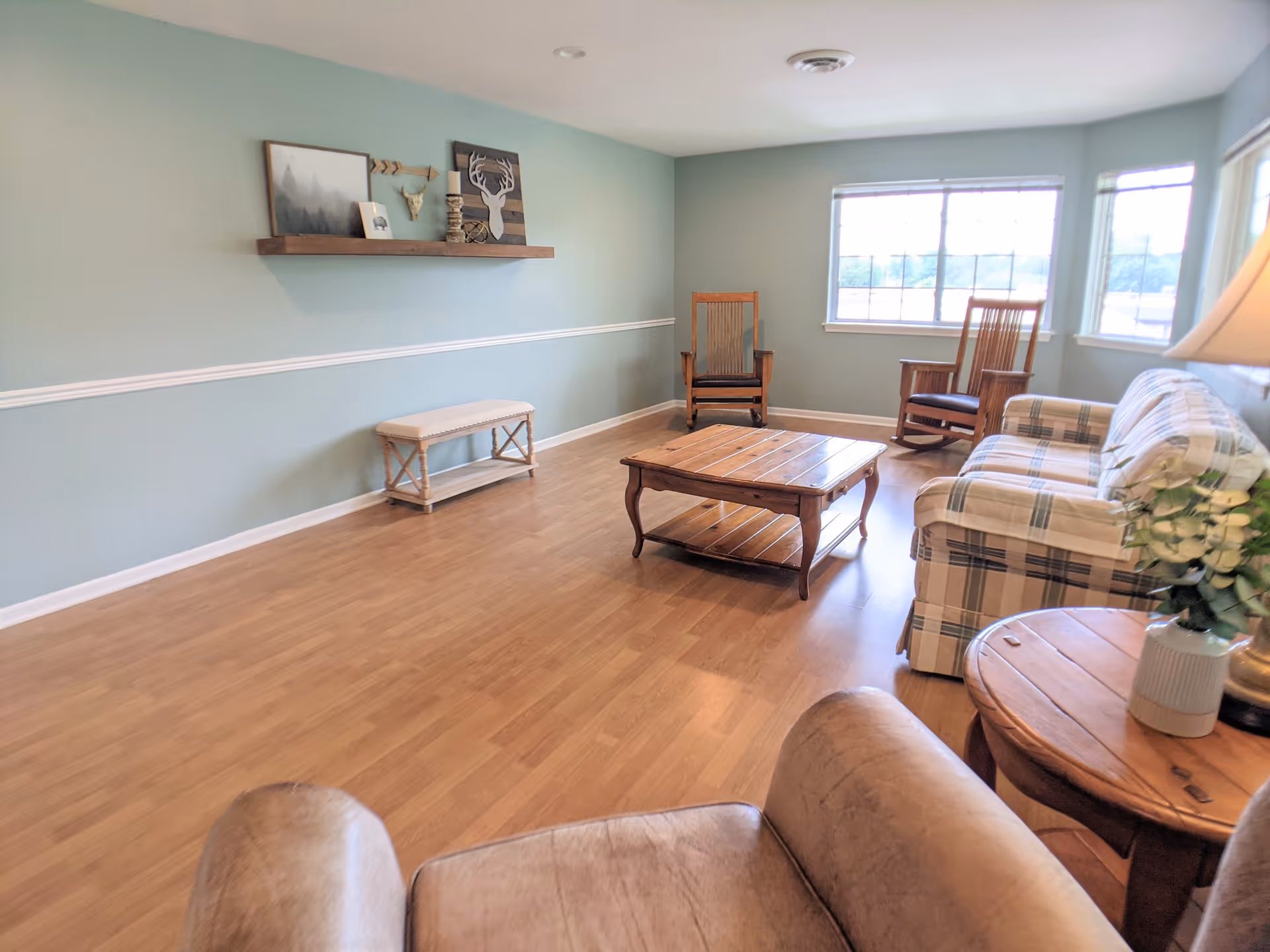 Bright sitting room with wood floors, a plaid sofa, armchairs, a wooden coffee table, and wall decor on a pale green wall.