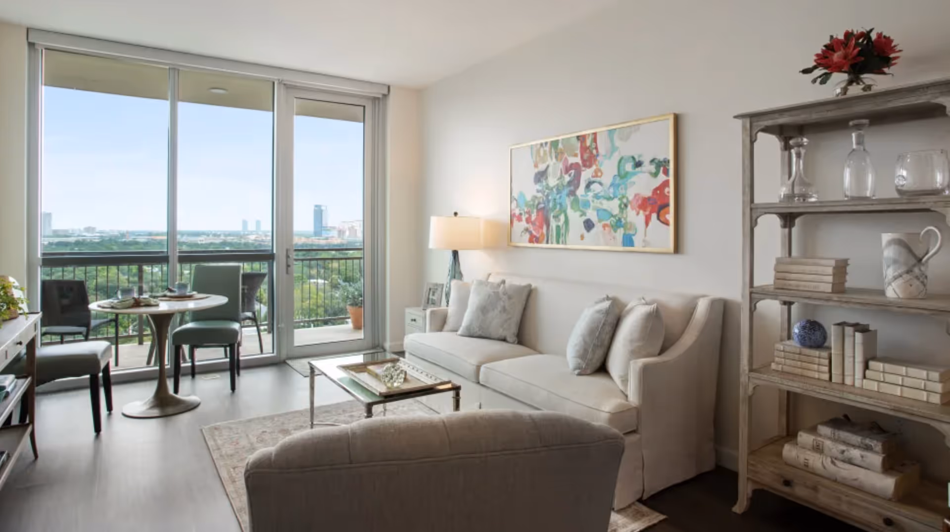 A bright living room with a large window and sliding glass door leading to a balcony with a city view. The room features a beige sofa with cushions, a glass coffee table, a small round dining table with two chairs, a floor lamp, a colorful abstract painting on the wall, and a wooden shelving unit with books, glassware, and a vase with red flowers.