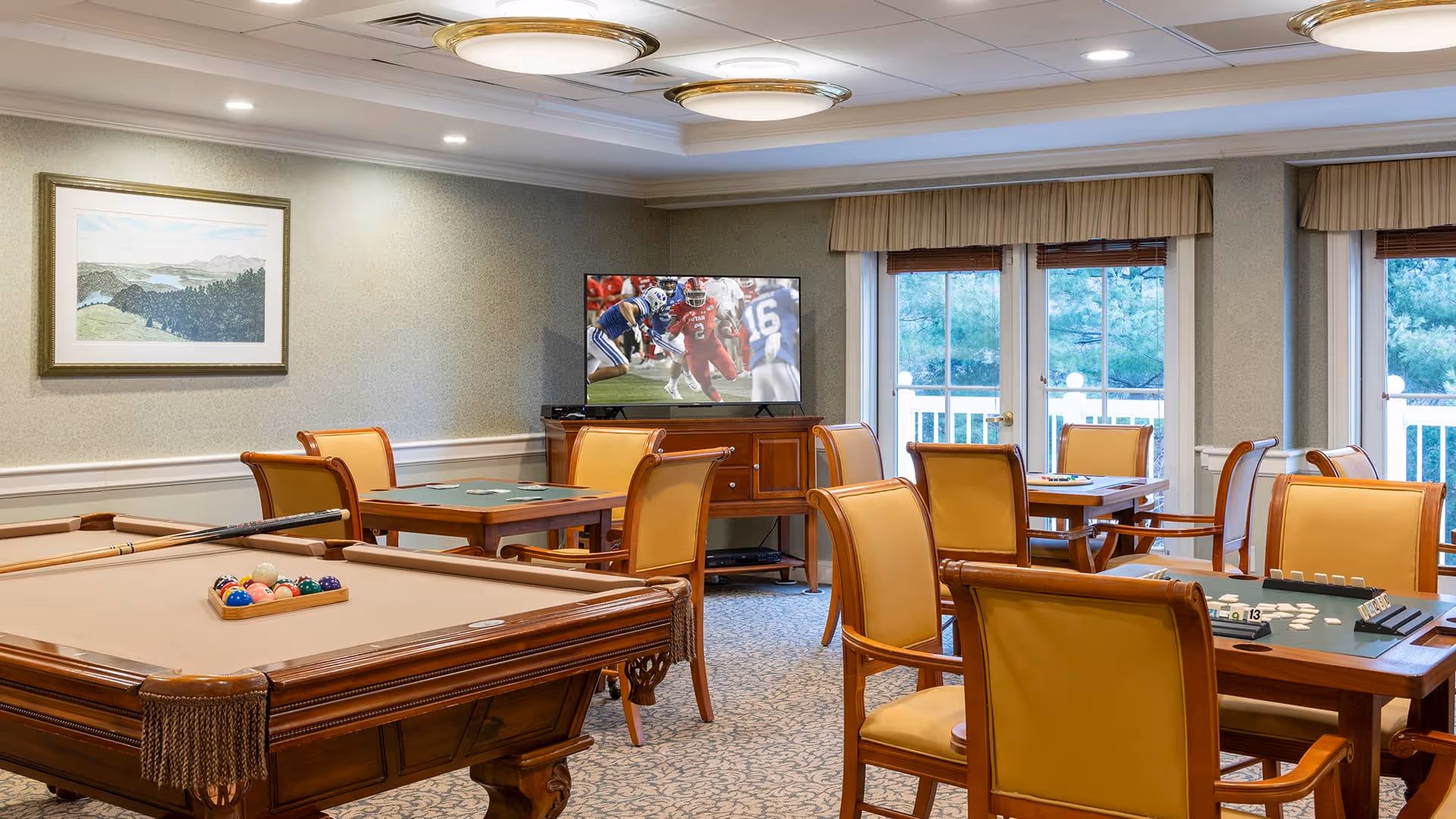 A well-lit recreational room with a pool table in the foreground, several tables with yellow cushioned chairs arranged for card or board games, a flat-screen TV showing a football game, large windows with beige valances letting in natural light, and a framed landscape painting on the wall.