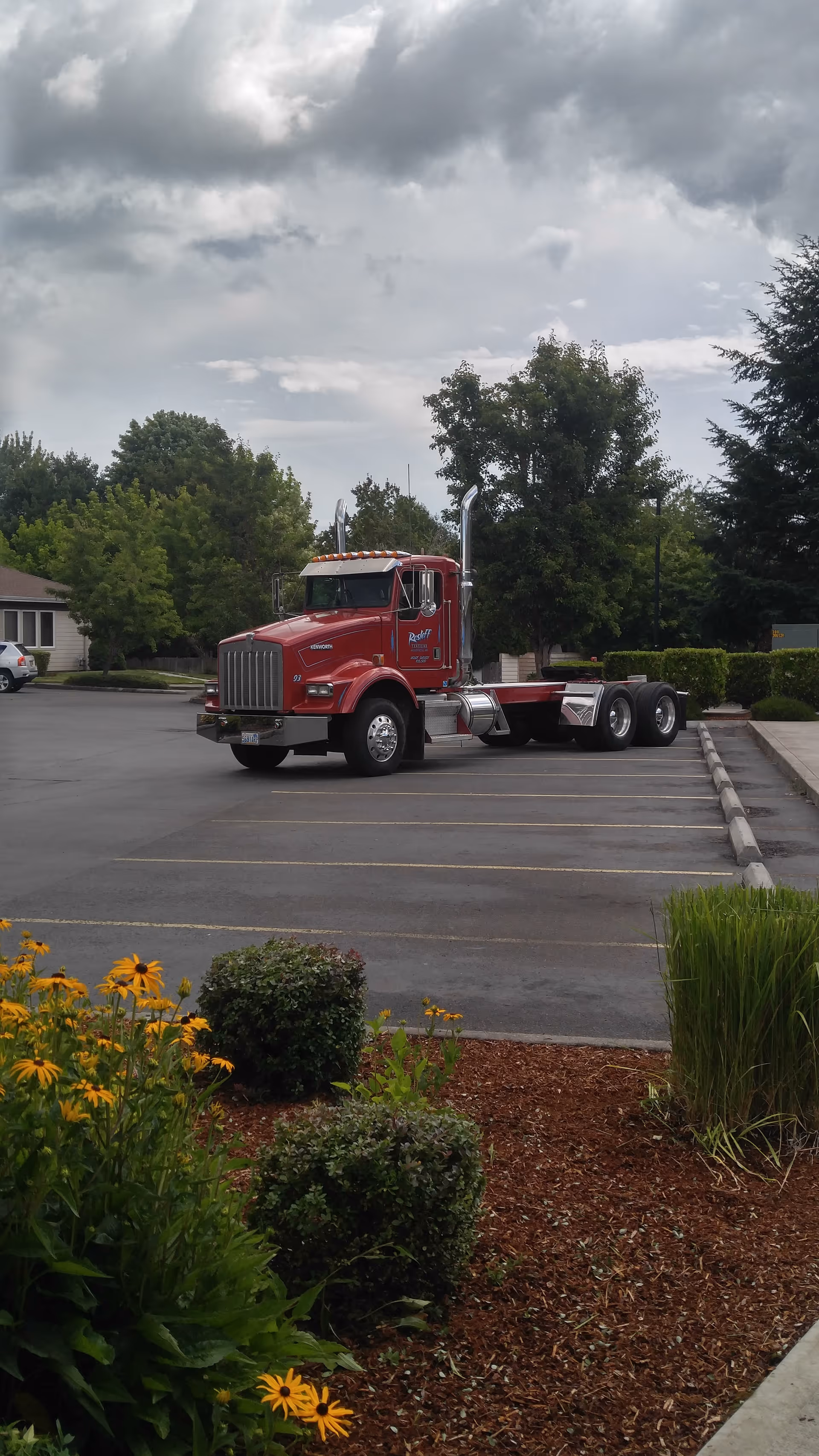 A red semi-truck parked in an empty parking lot with trees and bushes in the background under a cloudy sky. There are yellow flowers and green shrubs in the foreground near a mulched garden bed.