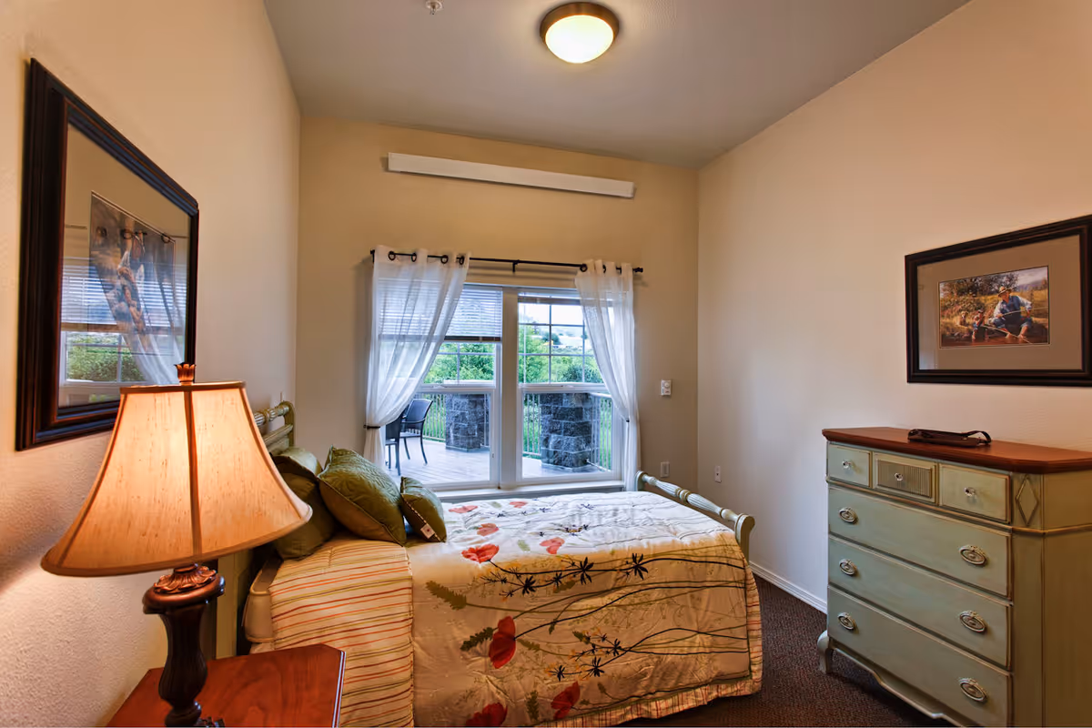A cozy bedroom in a senior living community featuring a bed with floral bedding, a bedside table with a lamp, a green dresser, framed artwork on the walls, and a window with sheer white curtains overlooking an outdoor patio with chairs.