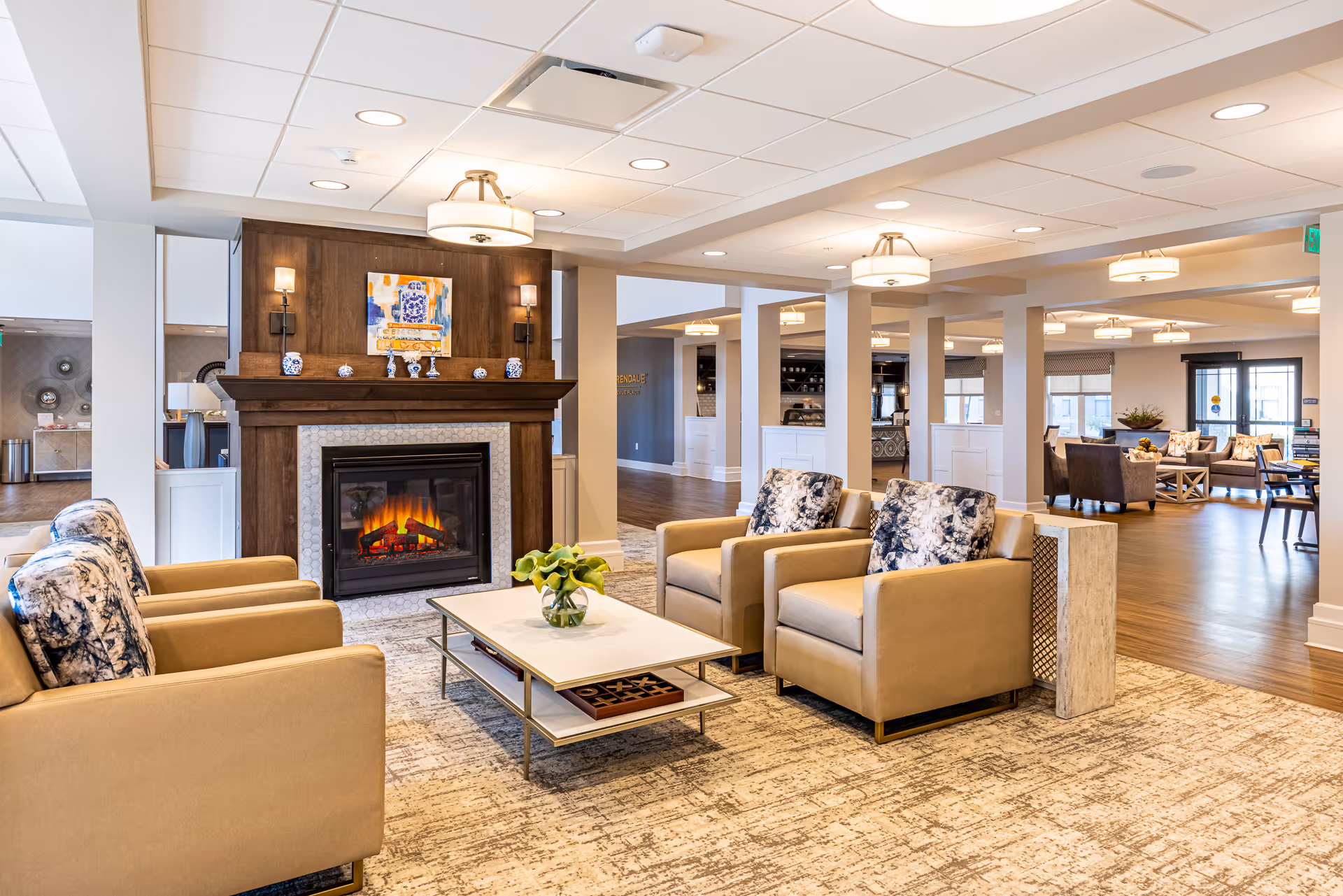 Bright open lounge with armchairs and a coffee table arranged around a lit fireplace in a senior living facility.