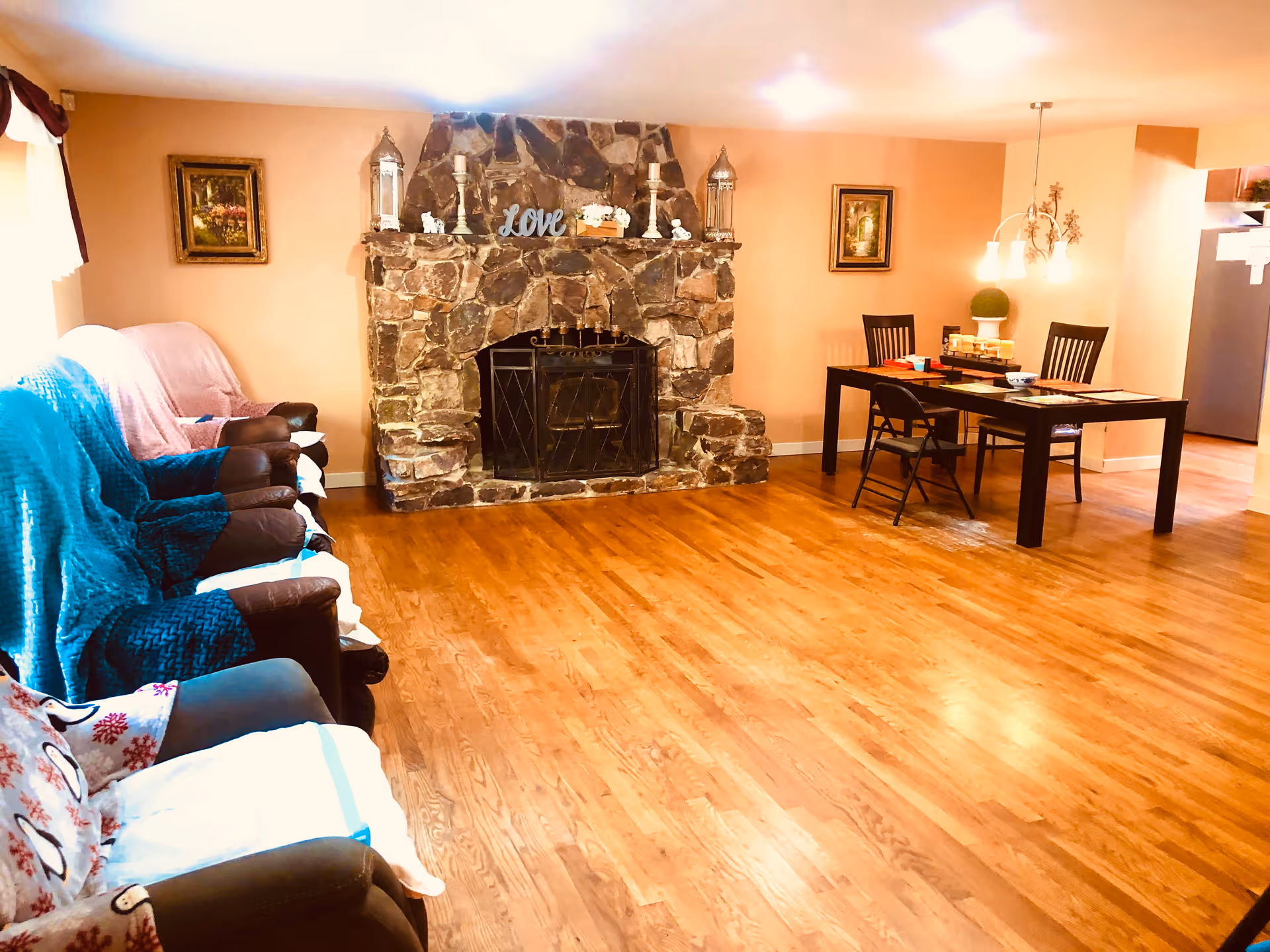 A cozy living room area with a stone fireplace in the center, decorated with candles, lanterns, and a 'Love' sign on the mantel. To the left, there are several armchairs covered with blankets. To the right, there is a dining table with chairs and a hanging light fixture above it. The room has wooden flooring and warm-colored walls with framed pictures.