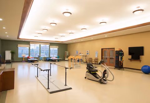 A spacious rehabilitation or physical therapy room with parallel bars for walking exercises, an exercise bike, therapy tables near large windows, chairs, a wall-mounted TV, and exercise equipment including a large blue exercise ball. The room has a high ceiling with recessed lighting and a warm color scheme.