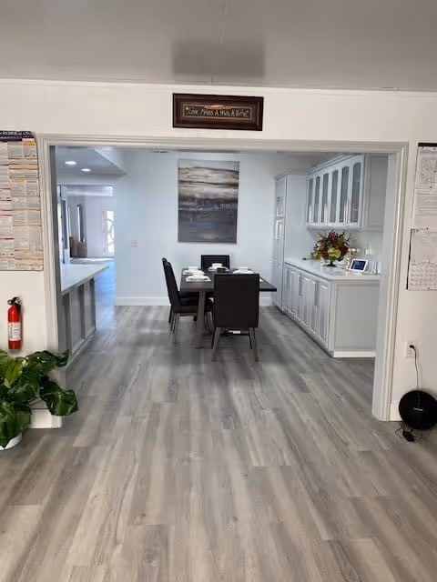 Interior view of a senior living facility showing a dining area with a table set for four and black chairs. To the right, there is a white cabinet with glass doors on the upper section and a flower arrangement on the countertop. The floor is light wood, and there is a large piece of artwork on the far wall. A fire extinguisher and a green plant are visible on the left side near the entrance.