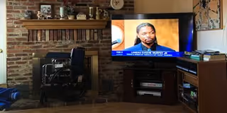 A cozy living room with a brick fireplace on the left, a flat-screen TV on a wooden stand showing a man speaking, and books and decorative items on shelves and mantle.