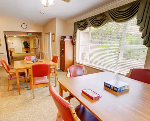 A bright dining room with wooden tables and red cushioned chairs. One table has a board game and a book on it. Large windows with green valance curtains let in natural light, and there is a hallway visible in the background with a clock and exit sign above the door.