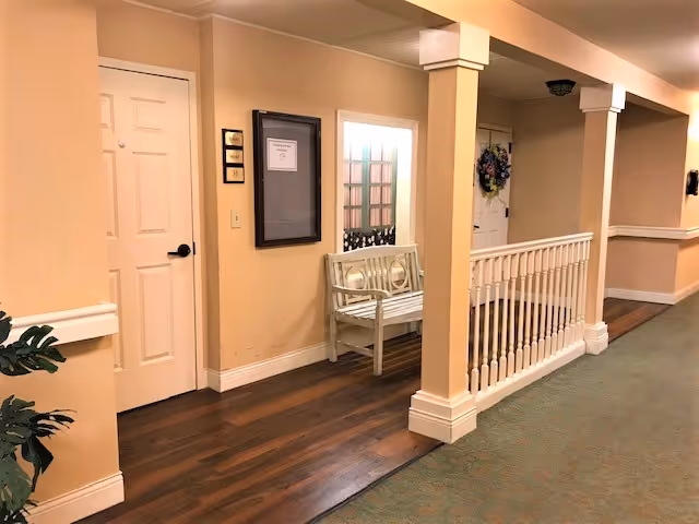 Interior hallway with beige walls, a white bench, railing and doors in a senior living facility.