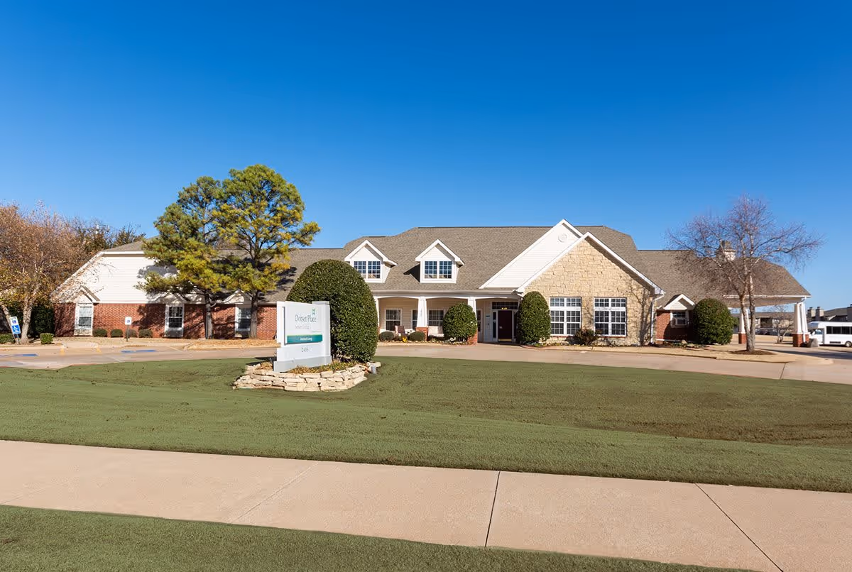 Exterior view of Dorset Place Memory Care facility showing a single-story building with a combination of brick and stone facade, a manicured lawn, bushes, trees, and a clear blue sky. A sign with the facility name is visible in front of the building.