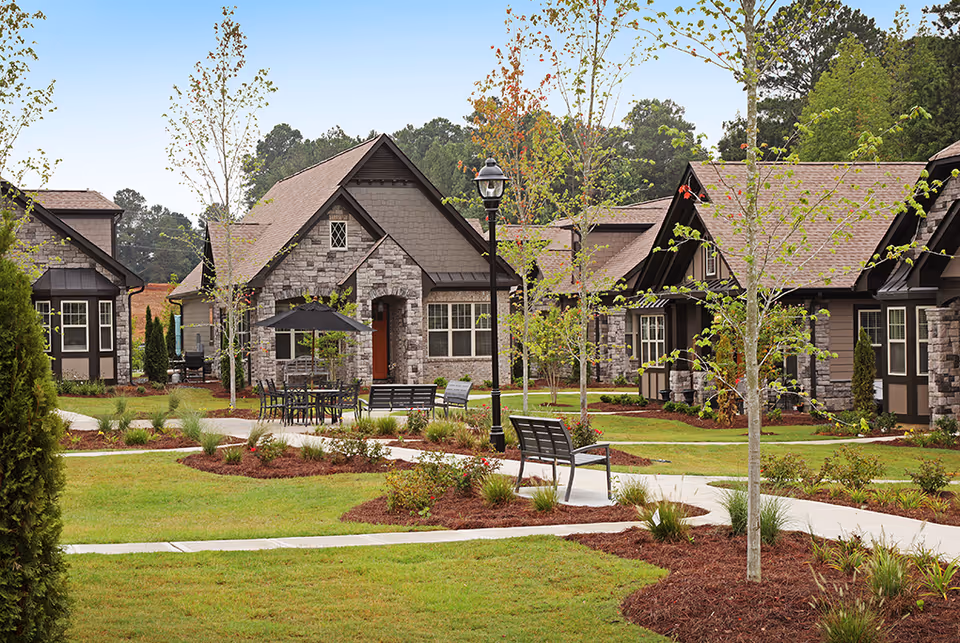 A landscaped outdoor area in a senior living community featuring stone and wood cottages with pitched roofs. There are young trees, green lawns, mulched flower beds, a paved walkway, benches, a table with chairs, and a black umbrella. A black lamppost stands near the center of the image.