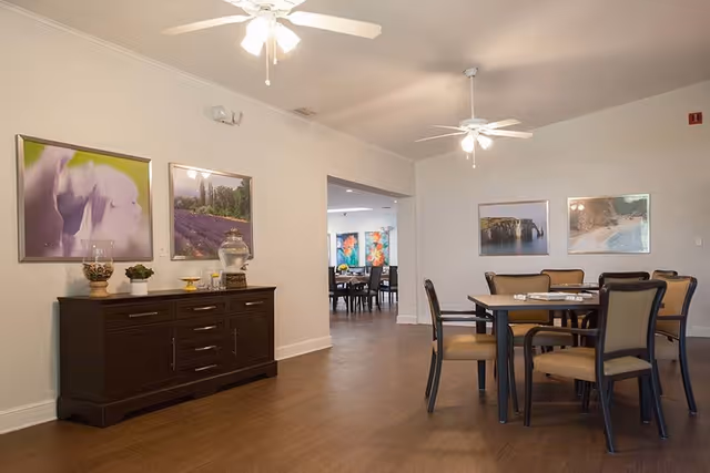 Interior view of a senior living facility dining area with a round table and six chairs. The room has wooden flooring, white walls, ceiling fans with lights, and framed landscape and floral artwork on the walls. A dark wooden sideboard with decorative items is positioned against one wall. Another dining area is visible through an open doorway.
