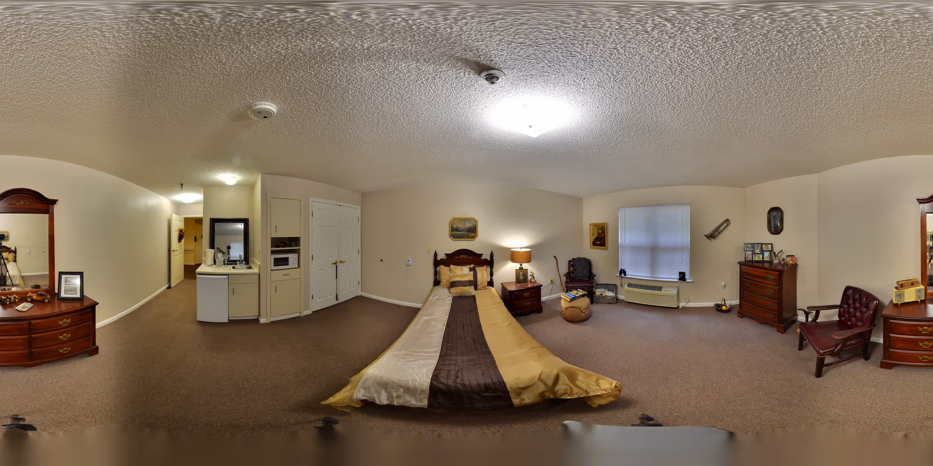 A spacious senior living bedroom with a single bed covered in a gold and brown bedspread. The room features beige walls and carpet, a wooden dresser with a mirror on the left, a small kitchenette area with a microwave and mini fridge, a nightstand with a lamp next to the bed, a comfortable chair with a footrest near a window with closed blinds, and another wooden dresser with framed photos and a chair on the right side.