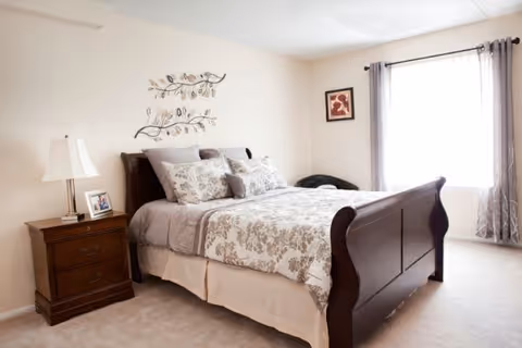 A cozy bedroom with a wooden sleigh bed dressed in floral patterned bedding and multiple pillows. There is a wooden nightstand with a lamp and a framed photo on it. The room has beige walls, a window with gray curtains, and a decorative metal wall art above the bed.