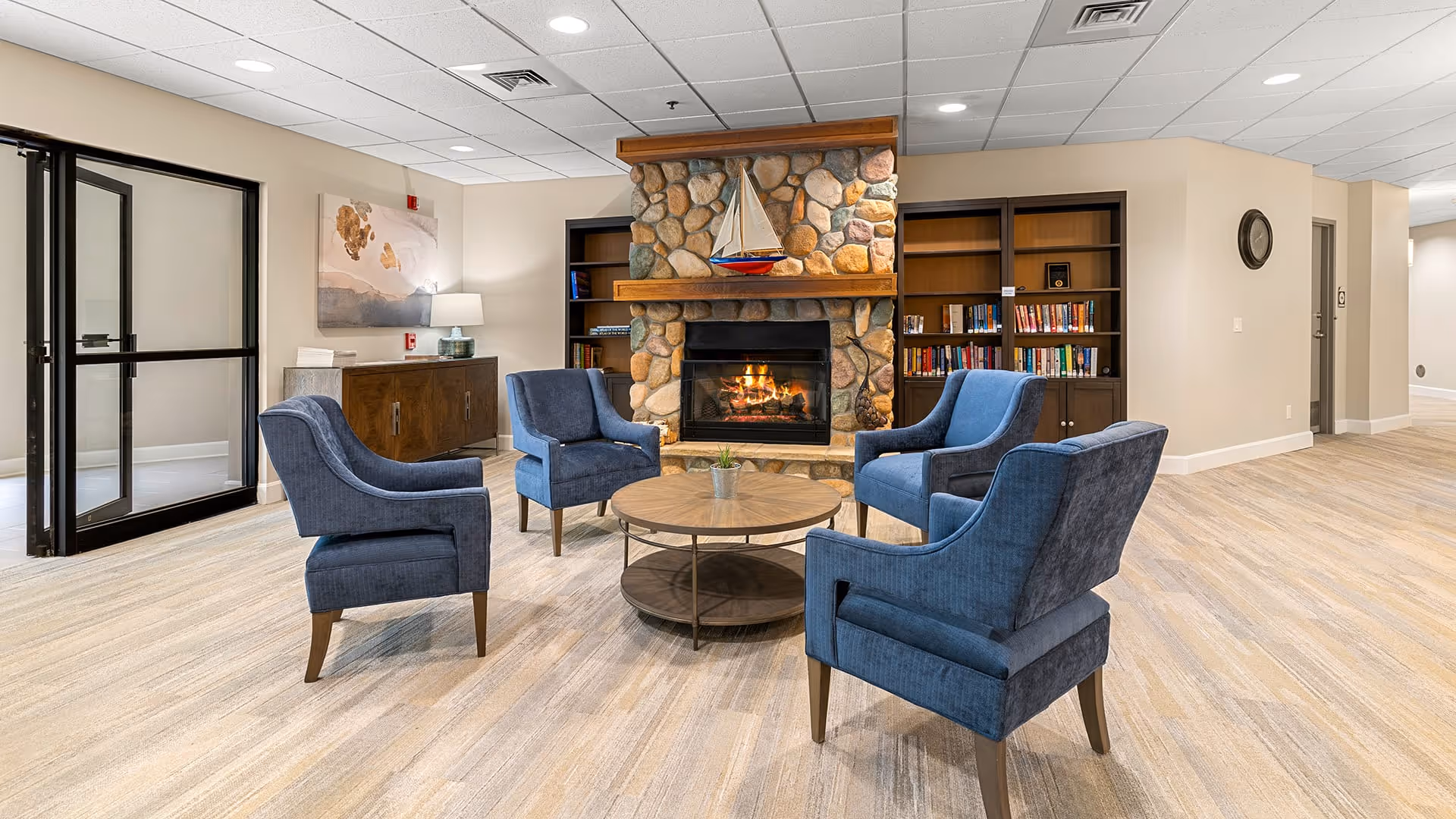 A cozy sitting area in a senior living facility with four blue upholstered armchairs arranged around a round wooden coffee table. Behind the seating is a stone fireplace with a model sailboat on the mantel and built-in wooden bookshelves filled with books on either side. The room has light-colored walls, a beige carpet, and recessed ceiling lights.