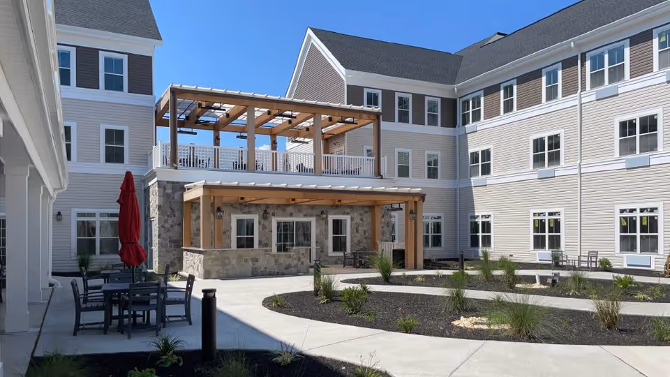 Outdoor courtyard area of a senior living facility with a paved walkway, landscaped garden beds, patio tables and chairs, a red closed umbrella, and a two-story building with beige siding and multiple windows under a clear blue sky.