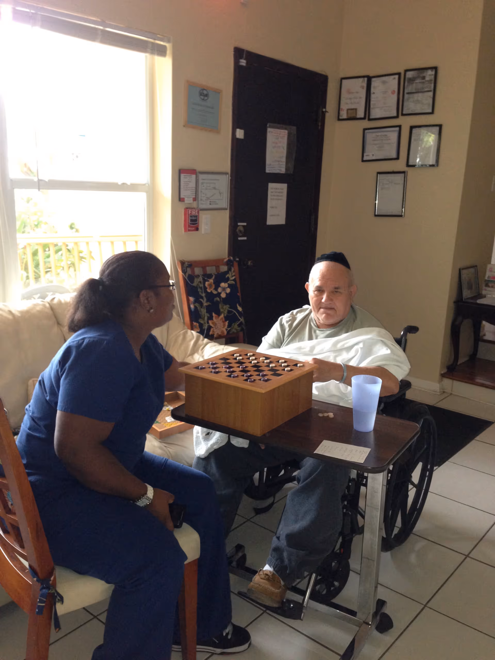 An elderly man in a wheelchair playing a board game with a caregiver in a blue uniform inside a room with certificates on the wall and a window letting in natural light.