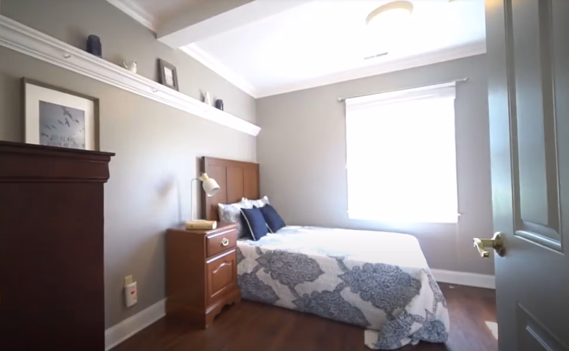 A small bedroom with a bed covered in a white and gray patterned comforter and several pillows. There is a wooden nightstand with a lamp and a book next to the bed. A wooden dresser is visible on the left side of the room. The walls are painted light gray, and there is a window letting in bright natural light. A shelf above the dresser holds decorative items and a framed picture.