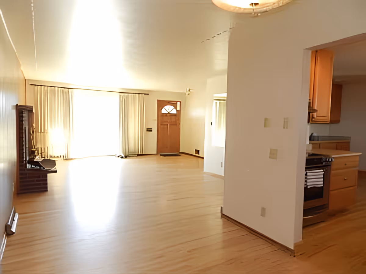 Bright open living room with hardwood floors, a front door and large curtained window, a fireplace on the left and a partial view into the kitchen on the right.