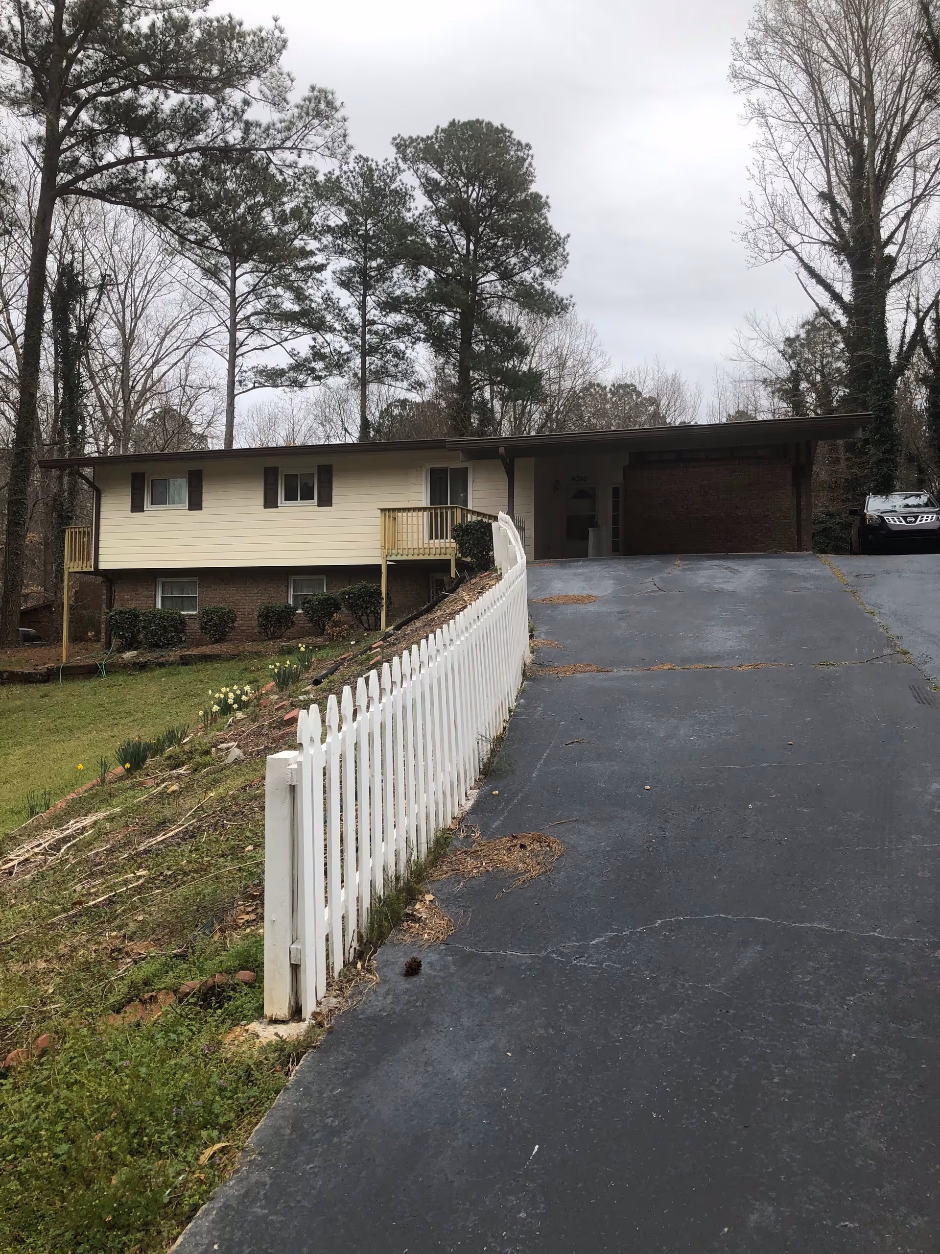 Front view of a single-story house with a sloped asphalt driveway, white picket fence, carport, and tall trees in the background.