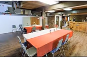 Interior view of a senior living facility dining area with a long table covered by a red tablecloth and surrounded by chairs. The room features wooden flooring, a bar counter in the background, and a ceiling with exposed beams and lighting fixtures.