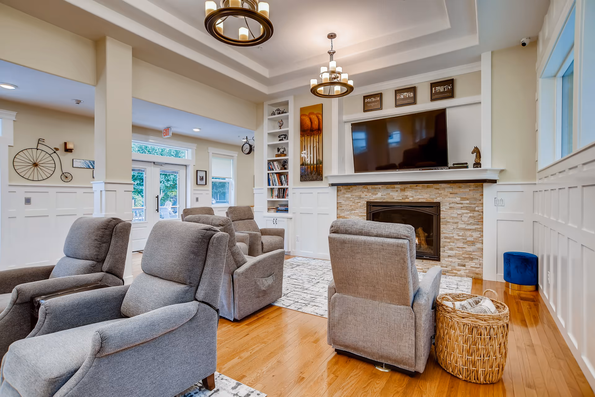 A cozy living room area with four gray upholstered recliner chairs arranged around a stone fireplace with a mounted flat-screen TV above it. The room features hardwood floors, white wainscoting on the walls, built-in shelves with books and decorative items, and two modern chandeliers hanging from a tray ceiling. There is a woven basket with a blanket next to one chair and a blue ottoman near the window. Large windows and glass doors provide natural light and a view of greenery outside.
