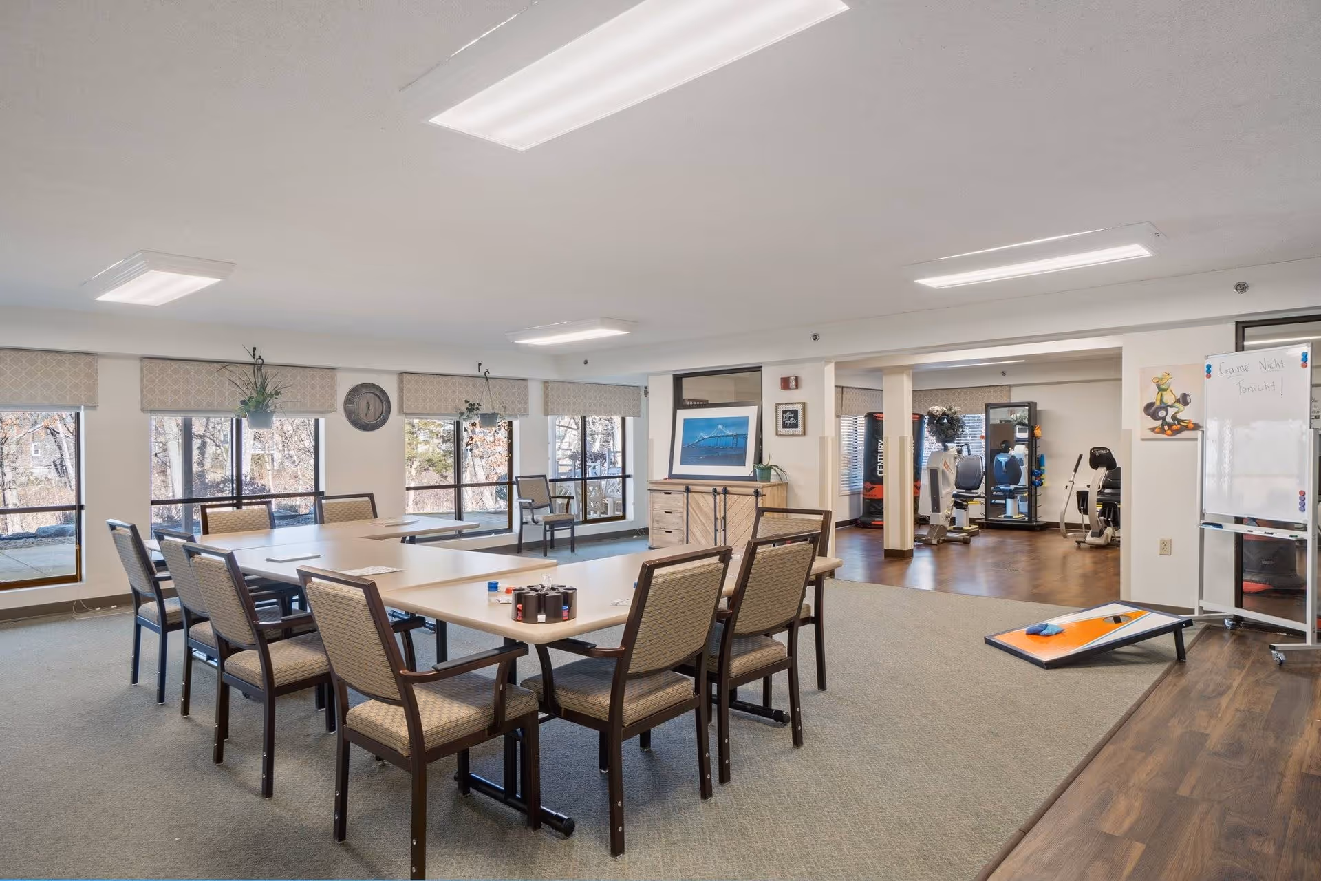 A spacious, well-lit common area with several tables and chairs arranged for group activities or dining. Large windows with beige valances let in natural light and offer a view of trees outside. In the background, there is an exercise area with fitness equipment and a whiteboard with a cartoon drawing and some writing. A cornhole game board is set up on the wooden floor near the exercise area.