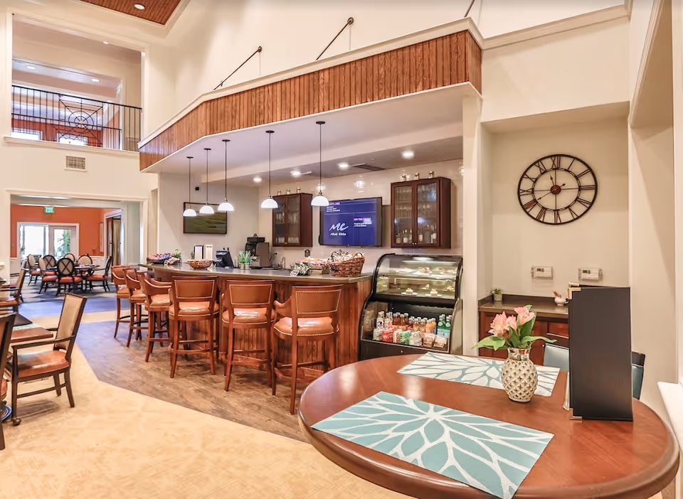 Interior view of a senior living facility's dining and snack bar area with wooden bar stools, pendant lights, a display fridge with beverages and snacks, a round table with placemats and a flower vase, and a large wall clock. In the background, there is a dining room with tables and chairs.