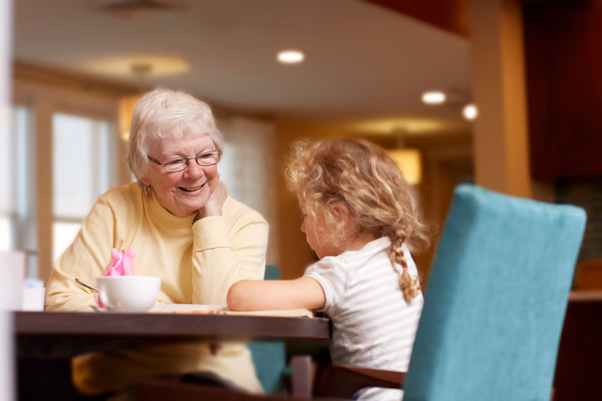 An elderly woman with glasses and a yellow sweater smiling and interacting with a young girl with curly hair sitting at a table in a warmly lit room.
