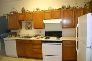 A kitchen with wooden cabinets, a white electric stove with four burners, a white refrigerator, a dishwasher, and a countertop with a sink. Decorative items and plants are placed on top of the cabinets.