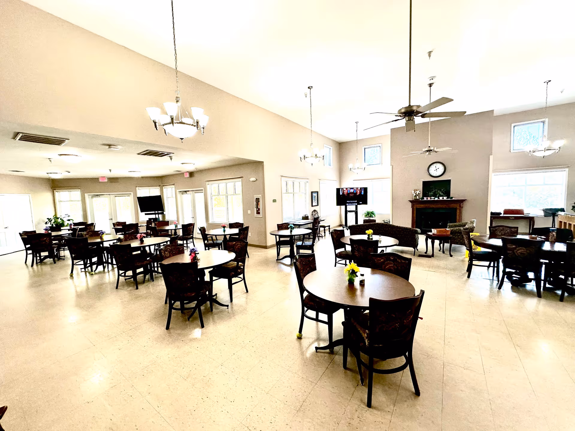 A spacious and well-lit dining room with multiple round and rectangular tables surrounded by chairs. Each table has a small flower arrangement. The room features large windows, ceiling fans, chandeliers, a fireplace with a clock above it, and a television mounted on a stand. The floor is tiled and the walls are painted in a light neutral color.