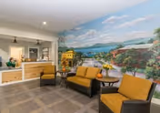 A cozy seating area in a memory care facility with three cushioned chairs and a small table, set against a large mural depicting a scenic landscape with trees, water, and mountains. In the background, there is a reception desk with a staff member behind it and a doorway leading to another room.