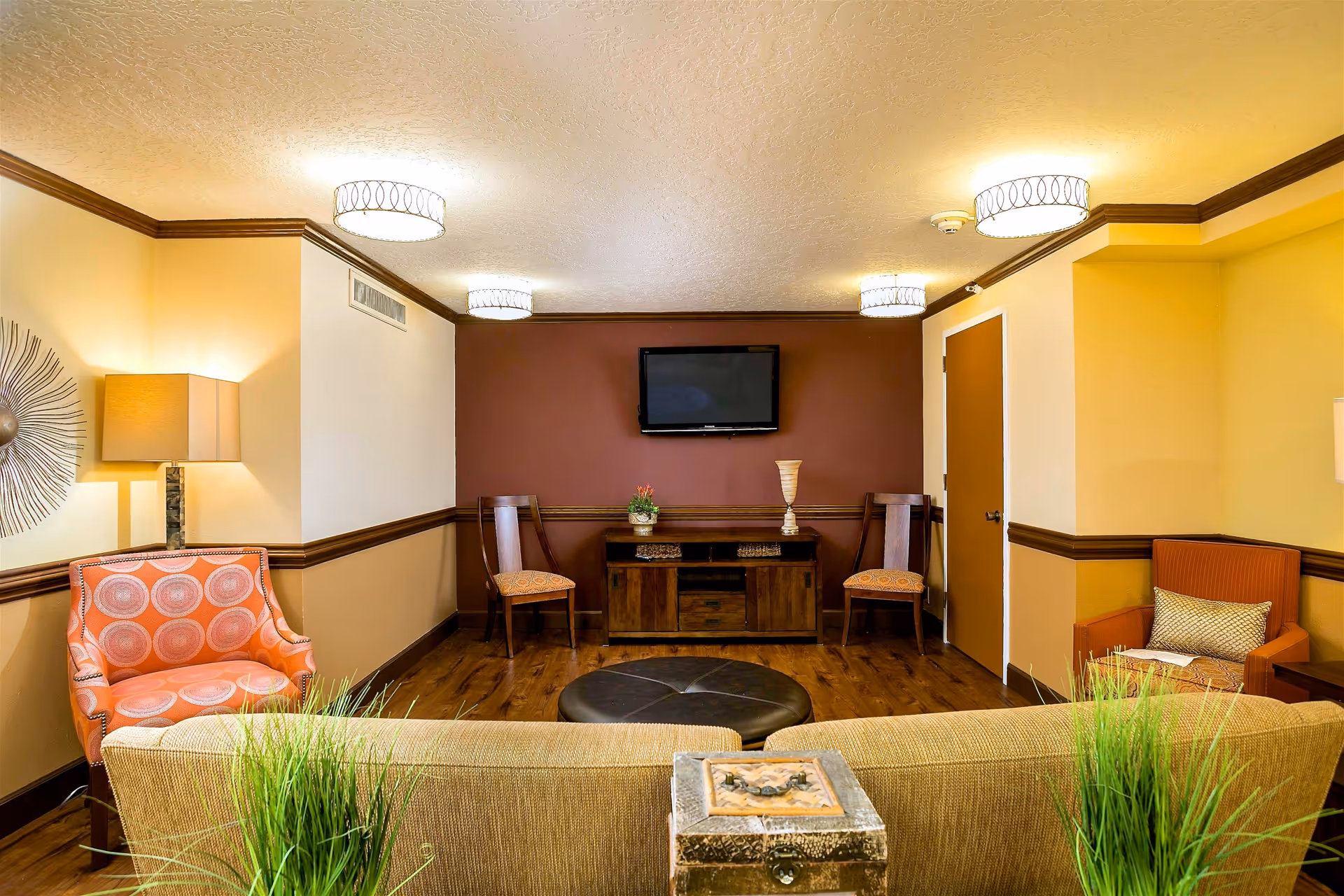A cozy living room area with a beige sofa in the foreground, a round black ottoman, two wooden chairs with patterned cushions, an orange armchair, and a small wooden cabinet with decorative items. A flat-screen TV is mounted on a maroon accent wall. The room features warm lighting from ceiling fixtures and a table lamp, with wood flooring and beige walls accented by dark brown trim.
