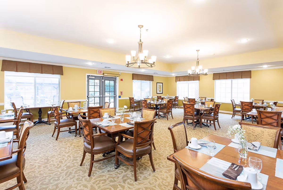 A bright and spacious dining room with multiple wooden tables and chairs arranged neatly. Each table is set with white cups, glasses, napkins, and small flower vases. The room has large windows with brown valances, beige walls, and carpeted floors with a subtle pattern. Two chandeliers hang from the ceiling, providing warm lighting.