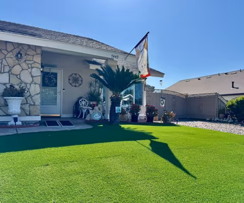 Front exterior view of a single-story house with a stone facade and a small porch area decorated with plants, chairs, and a flag. The lawn is green and well-maintained under a clear blue sky.