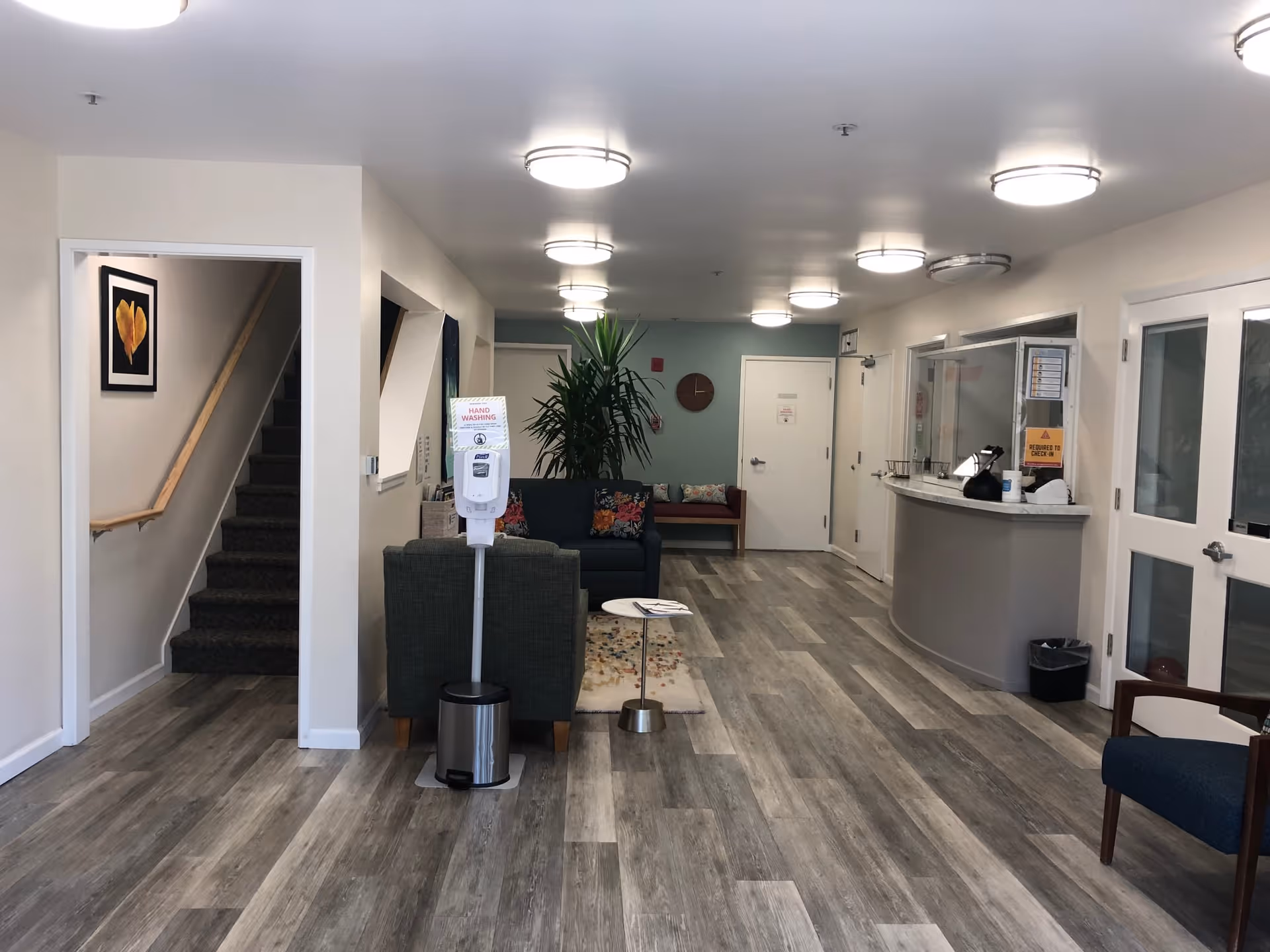 A clean and well-lit interior common area with wood-patterned flooring, a hand sanitizer station, seating with chairs and a sofa, a small table, a plant, a clock on the wall, and a reception desk with glass windows. There is a staircase on the left side and multiple doors along the right wall.