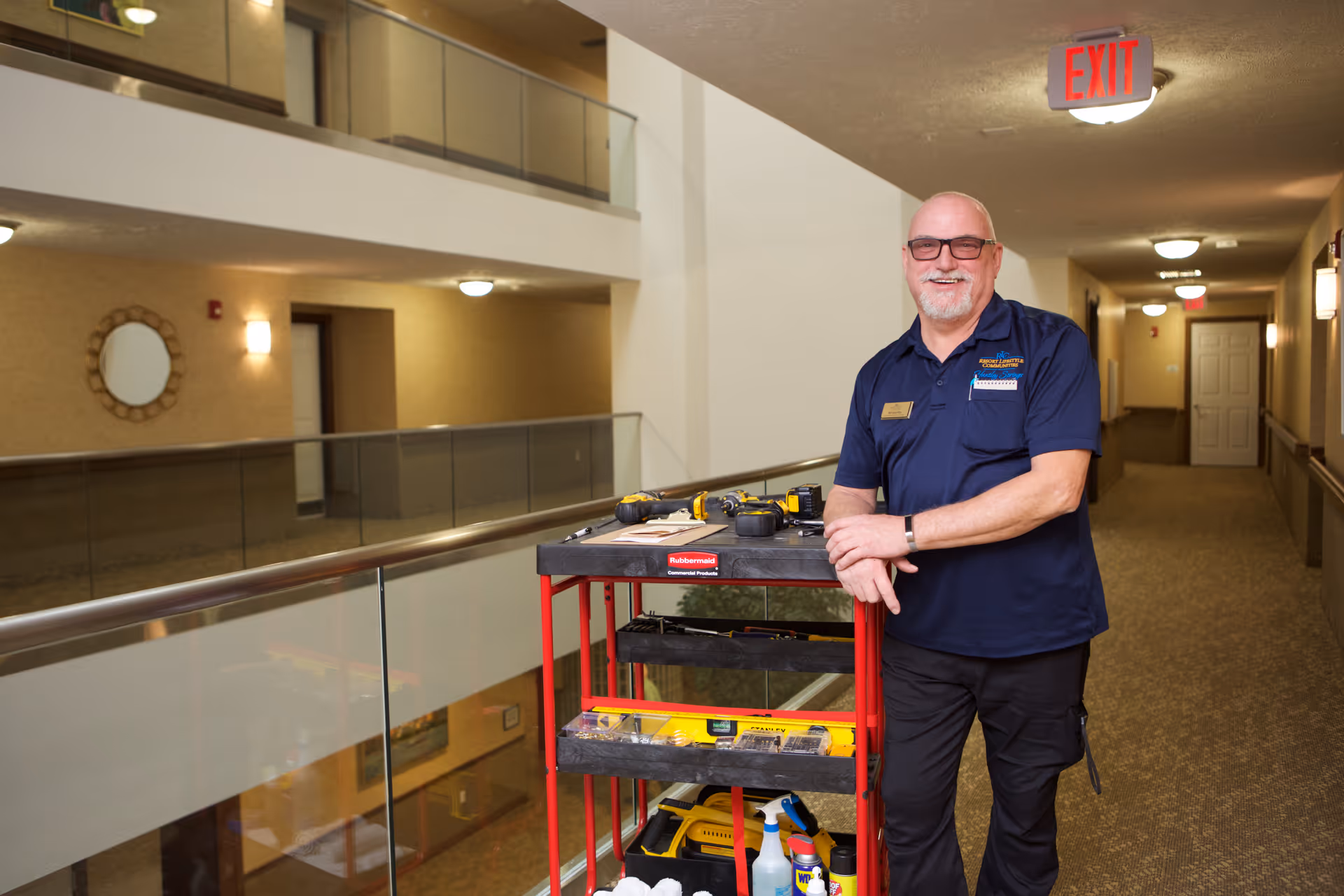 A smiling maintenance worker wearing glasses and a navy blue shirt stands in a hallway of a retirement facility next to a red utility cart filled with tools and cleaning supplies. The hallway has beige walls, carpeted floors, and glass railings overlooking a lower level. An exit sign is visible on the ceiling.