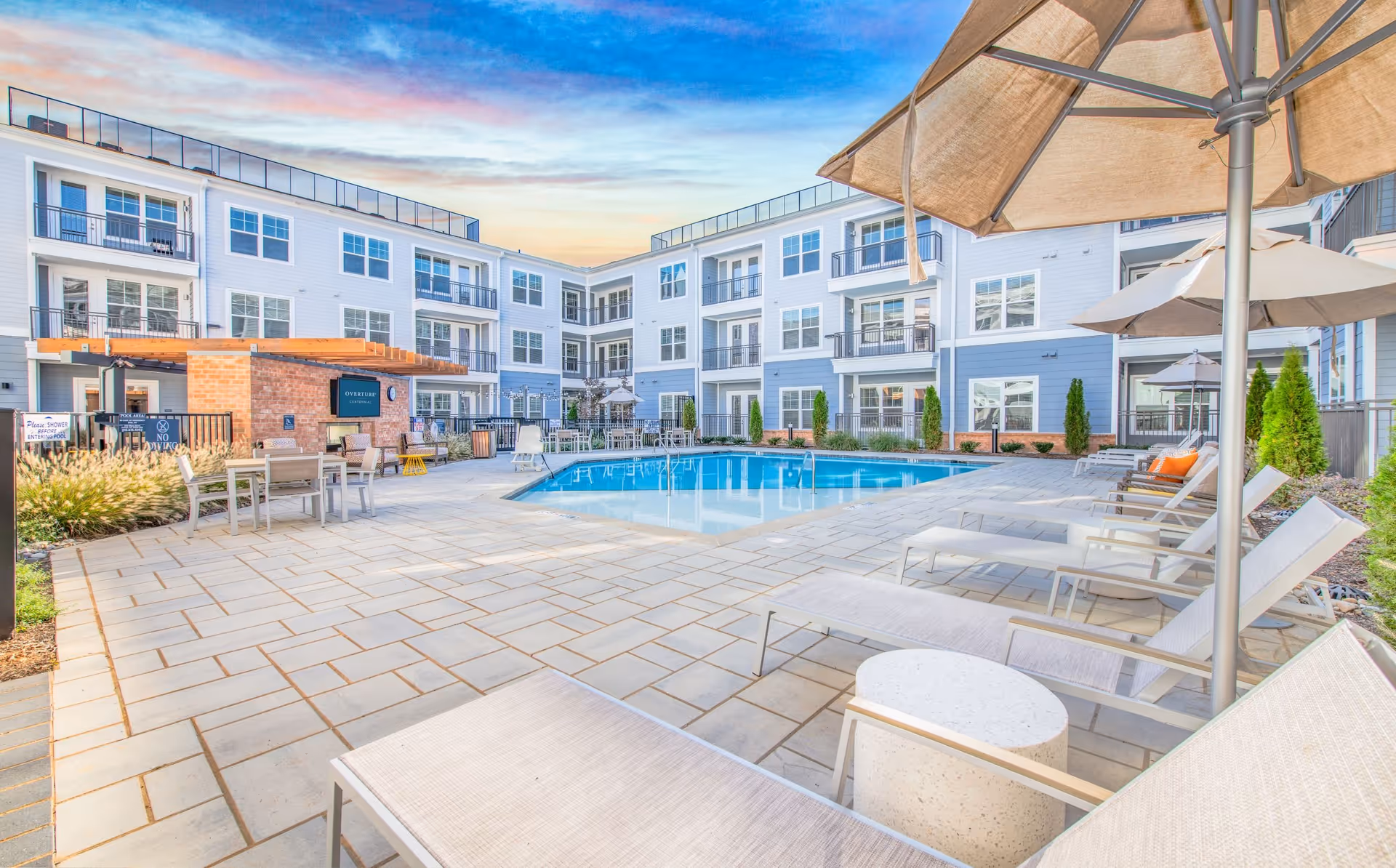 Outdoor pool area at Overture Centennial with lounge chairs, umbrellas, tables, and a three-story residential building surrounding the pool under a colorful sky at sunset.