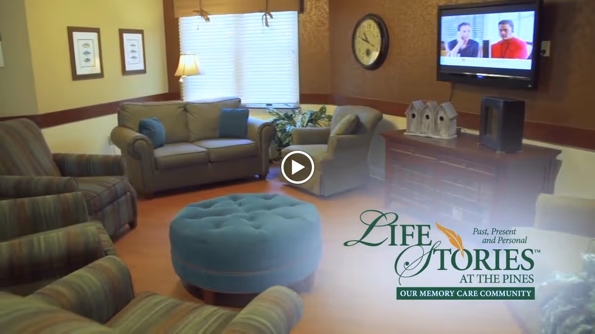 A cozy living room area with multiple upholstered chairs and a sofa arranged around a large round blue ottoman. There is a window with blinds and a lamp in the corner, framed pictures on the wall, a wall clock, and a flat-screen TV mounted on the wall showing two people. A wooden cabinet below the TV holds decorative birdhouses and other items.