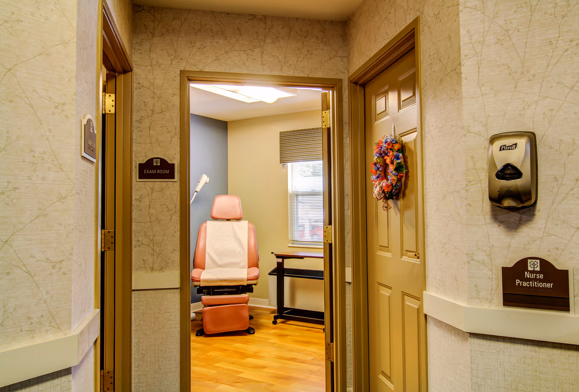 Hallway view into a medical exam room showing a reclining exam chair, hand sanitizer dispenser, and signs for an exam room and nurse practitioner.