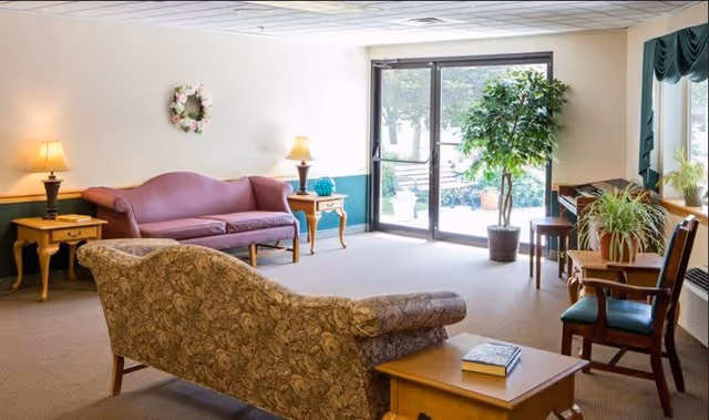 A bright senior living facility common area with a floral patterned sofa in the foreground, a purple couch against the wall, two wooden side tables with lamps, a small table with chairs near a window, and a large potted plant next to glass doors leading outside.