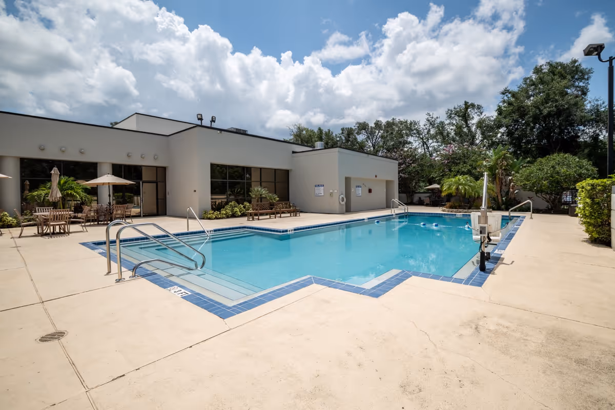Outdoor swimming pool area at Grand Villa of DeLand with clear blue water, poolside chairs and tables with umbrellas, surrounded by greenery and a modern building under a partly cloudy sky.