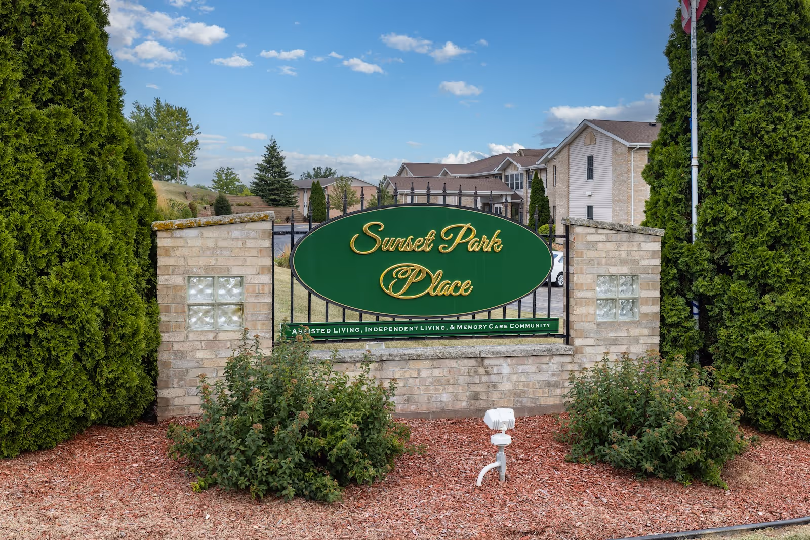 Entrance sign for Sunset Park Place, an assisted living, independent living, and memory care community, set between two brick pillars with greenery and a building in the background under a partly cloudy sky.