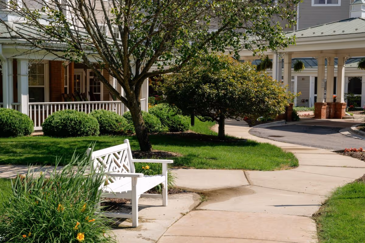 White bench by a curved walkway leading to the landscaped entrance and porch of an assisted living building.