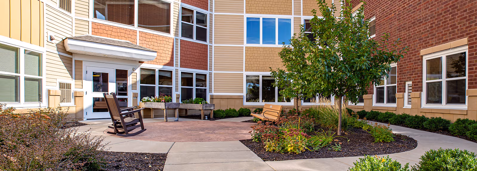 Outdoor courtyard area of a senior living facility with a curved concrete walkway, a small tree, various plants and shrubs, a wooden rocking chair, a bench, and raised garden beds in front of a multi-story building with large windows.