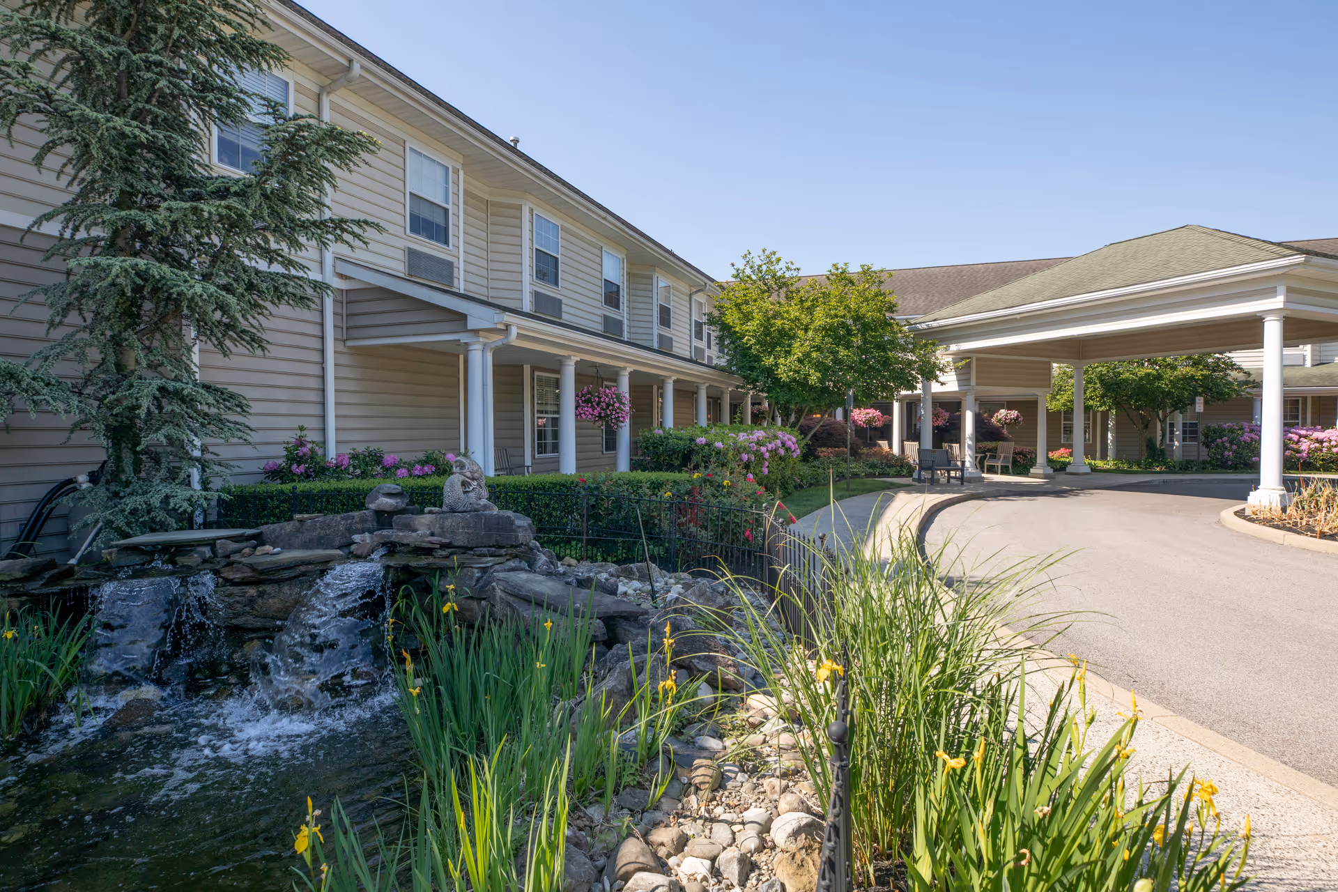 Landscaped front entrance of a senior living facility with a covered porte-cochere, a small pond and flowering plants.
