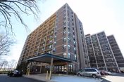 Front view of a multi-story brick senior living tower with a covered driveway entrance and parked cars.