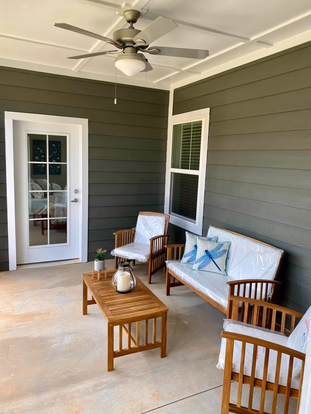 Covered outdoor patio area with green siding walls, a ceiling fan, a wooden bench and two wooden chairs with white cushions, a wooden coffee table with a candle and small plant, and a glass door leading inside.