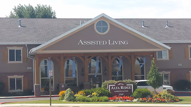 Front exterior view of Alta Ridge Assisted Living of Sandy, showing a two-story building with a peaked roof and a covered entrance. There are windows with wooden shutters, a landscaped garden with colorful flowers and shrubs, and a sign in front that reads 'Alta Ridge Assisted Living 1360'.