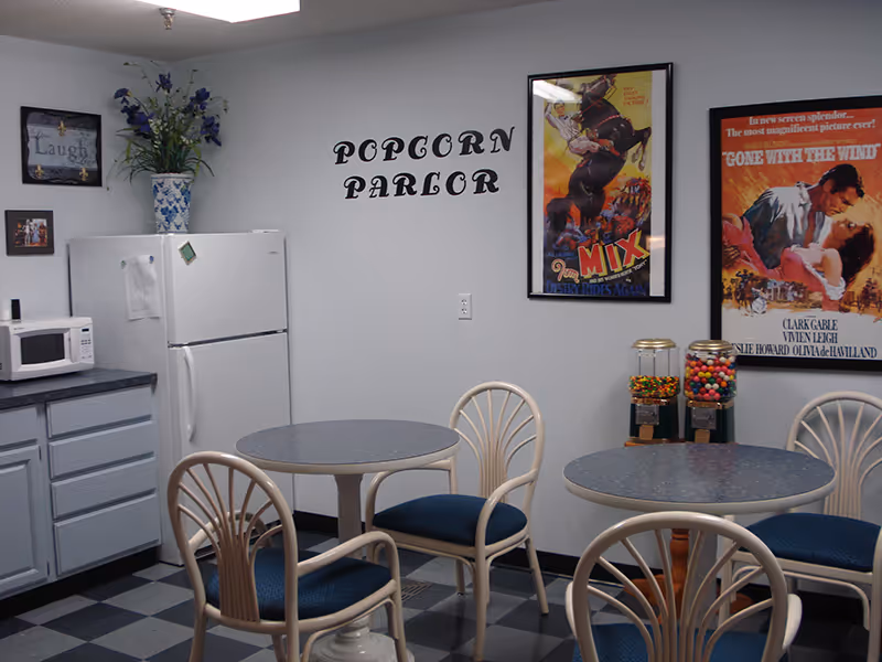 A small communal dining area labeled 'Popcorn Parlor' with round tables, blue-cushioned chairs, a microwave, a refrigerator, and framed posters on the wall.
