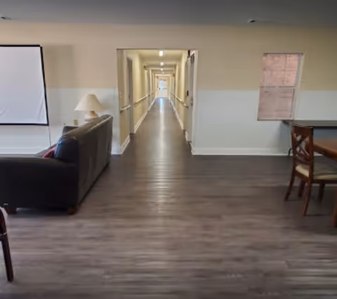 Interior view of a senior living facility showing a long hallway with handrails on both sides. In the foreground, there is a living area with a dark sofa, a lamp on a side table, and a dining table with chairs. The walls are painted beige with a white lower half, and the floor is covered with wood-like flooring.